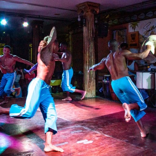 Homens dançam a Puxada de Rede em uma apresentação folclórica no palco, vestindo calças azuis, enquanto recriam a tradição baiana da pesca em uma performance animada.