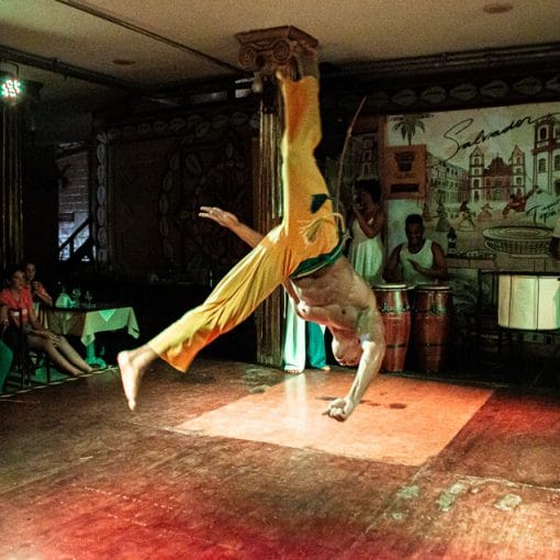 Capoeirista realiza um movimento acrobático no palco, exibindo a força e a agilidade típicas da capoeira, enquanto músicos tocam berimbau ao fundo no Show Folclórico de Salvador.