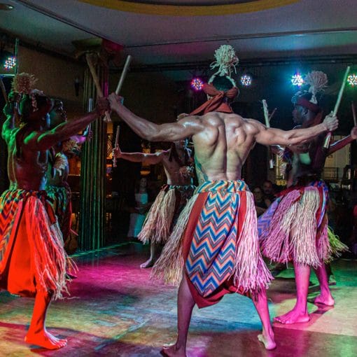 Dançarinos vestidos com saias de palha e trajes tradicionais realizam a dança do Maculelê, uma arte marcial afro-brasileira, batendo bastões em um palco iluminado.