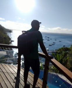 Guia turístico Jorge Oliveira, da De Boa Turismo, com mochila, observa a vista do mar e barcos de um mirante em Morro de São Paulo, Bahia, sob o sol.