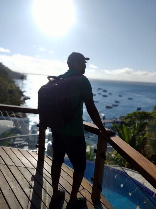 Guia turístico Jorge Oliveira, da De Boa Turismo, com mochila, observa a vista do mar e barcos de um mirante em Morro de São Paulo, Bahia, sob o sol.