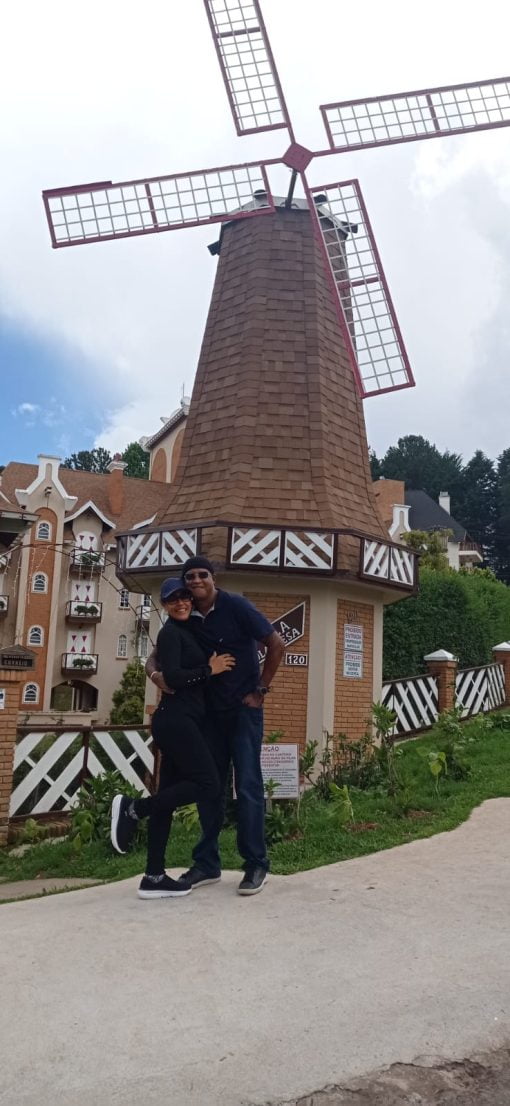 Casal feliz posando em frente a um moinho de vento em Campos do Jordão, com arquitetura de estilo europeu ao fundo.