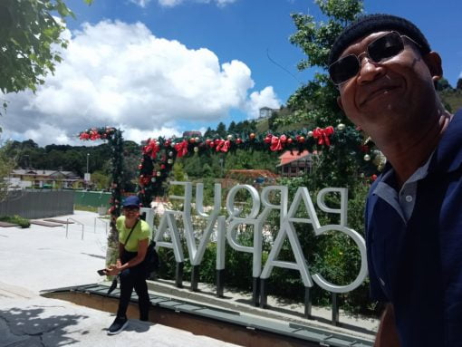 Casal sorridente posando em frente à entrada decorada do Parque Capivari em Campos do Jordão, em um dia ensolarado.