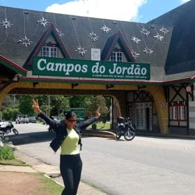 Turista sorridente posando em frente ao portal de entrada de Campos do Jordão, com decoração temática e bonecos de soldados.