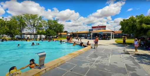 Pessoas relaxando em uma piscina de águas termais em Caldas do Jorro, Bahia, com um céu azul, árvores ao fundo e instalações ao redor.