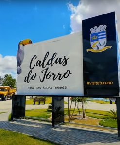 Placa de boas-vindas em Caldas do Jorro, Bahia, destacando o título "Terra das Águas Termais" com um céu azul e palmeiras ao fundo.