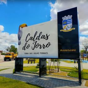 Placa de boas-vindas em Caldas do Jorro, Bahia, destacando o título "Terra das Águas Termais" com um céu azul e palmeiras ao fundo.