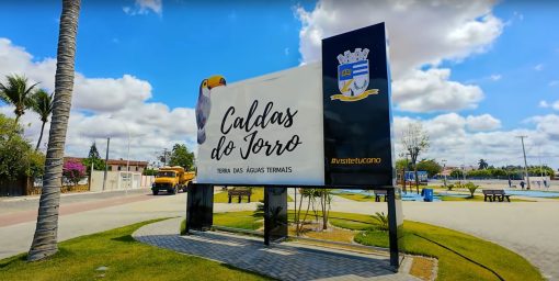 Placa de boas-vindas em Caldas do Jorro, Bahia, destacando o título "Terra das Águas Termais" com um céu azul e palmeiras ao fundo.