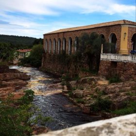 Imagem de um prédio histórico de arquitetura colonial, com arcadas e paredes de pedra, situado às margens de um rio com corredeiras e rochas, cercado por vegetação nativa. O céu azul com nuvens brancas complementa a paisagem encantadora de Lençóis, Chapada Diamantina.