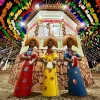 Estátuas estilizadas de três mulheres tocando instrumentos típicos do forró, vestidas com trajes coloridos e chapéus de cangaceiro. Ao fundo, um coreto decorado com bandeirinhas multicoloridas e elementos juninos, criando um cenário vibrante e festivo no São João de Lençóis, Chapada Diamantina.