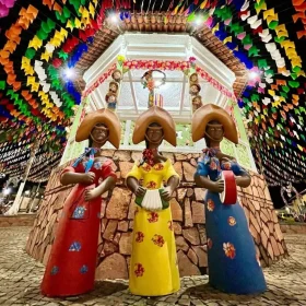Estátuas estilizadas de três mulheres tocando instrumentos típicos do forró, vestidas com trajes coloridos e chapéus de cangaceiro. Ao fundo, um coreto decorado com bandeirinhas multicoloridas e elementos juninos, criando um cenário vibrante e festivo no São João de Lençóis, Chapada Diamantina.